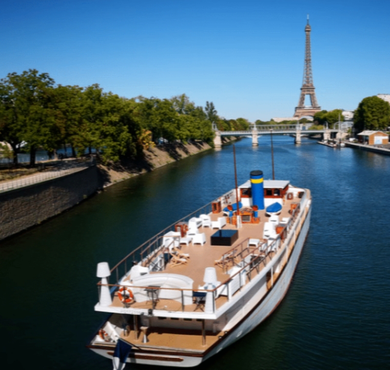 LE YACHT CLIPPER (75015) navigue sur la Seine à Paris, avec la Tour Eiffel et des arbres en arrière-plan sous un ciel bleu clair.
