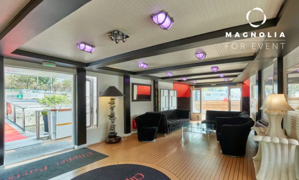 Salon moderne sur LE YACHT CLIPPER (75015) avec des canapés noirs, des tables en verre, des plafonniers violets et une lampe unique. De grandes fenêtres remplissent l'espace de lumière naturelle. Le logo de Magnolia For Event est affiché dans le coin supérieur droit.