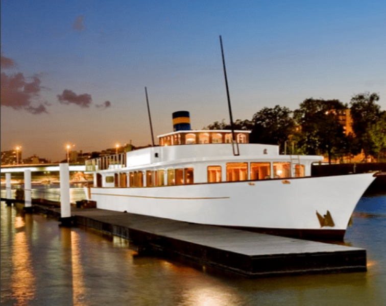 LE YACHT CLIPPER (75015), un grand yacht blanc avec un entonnoir bleu et jaune, est à quai au coucher du soleil, illuminé sur une eau calme qui reflète les lumières de la ville, avec des arbres et des bâtiments en arrière-plan.