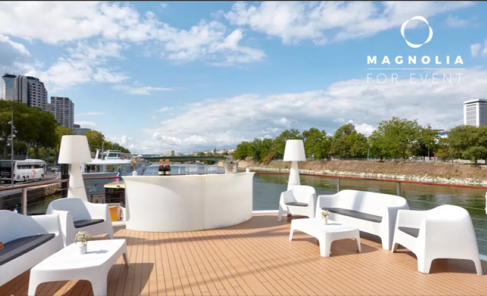 Une terrasse extérieure moderne sur LE YACHT CLIPPER (75015) comprend des canapés, des chaises et des tables blanches avec des lampes et des accents floraux, surplombant une rivière calme et des arbres luxuriants sous un ciel partiellement nuageux. Un magnolia pour l'événement est noté dans le coin.