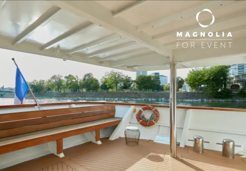 Vue du pont couvert de LE YACHT CLIPPER (75015) avec des bancs en bois, un drapeau français, une bouée de sauvetage et une rive pittoresque avec des arbres et des bâtiments en arrière-plan. Le logo "Magnolia for Event" apparaît dans le coin supérieur droit.