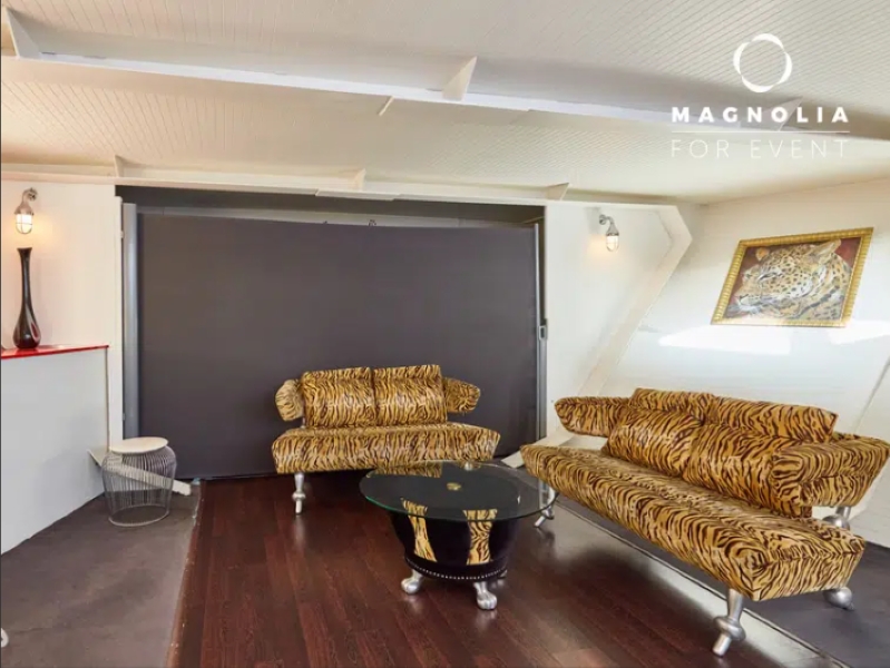 Salon moderne à bord du LE YACHT CLIPPER (75015) comprenant deux canapés imprimés tigre, une table basse ronde en verre, des planchers en bois foncé et un mur orné d'une grande photo de léopard. Le logo de Magnolia for Event est affiché dans le coin supérieur droit.