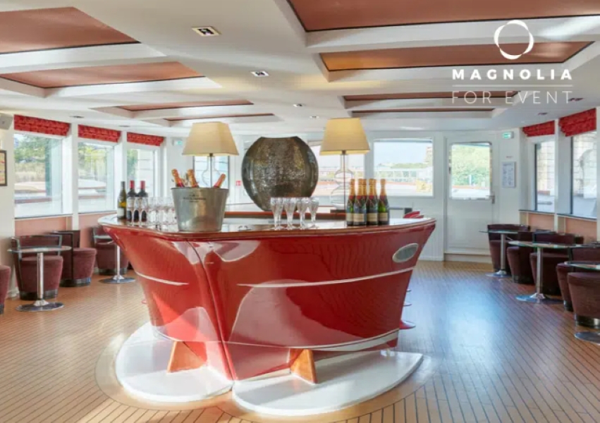 LE YACHT CLIPPER (75015) est un espace événementiel moderne doté d'un bar rouge brillant entouré de bouteilles de vin, de verres, de tabourets et de grandes fenêtres. La salle lumineuse est dotée d'un sol en bois, de deux lampes et d'un vase décoratif au-dessus du bar.