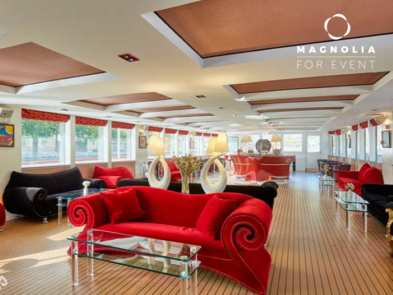 Une salle d'événement spacieuse et élégante à LE YACHT CLIPPER (75015) comprend des canapés en velours rouge et noir, des tables basses en verre, un plancher en bois, de grandes fenêtres et des lampes décoratives. Le logo Magnolia For Event apparaît dans le coin supérieur droit.