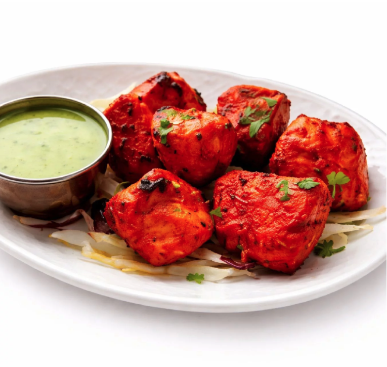 POULET TANDOORI (300g) : Morceaux de poulet tandoori grillés, garnis de coriandre et d'oignons émincés, servis dans une assiette ovale blanche avec un chutney vert.