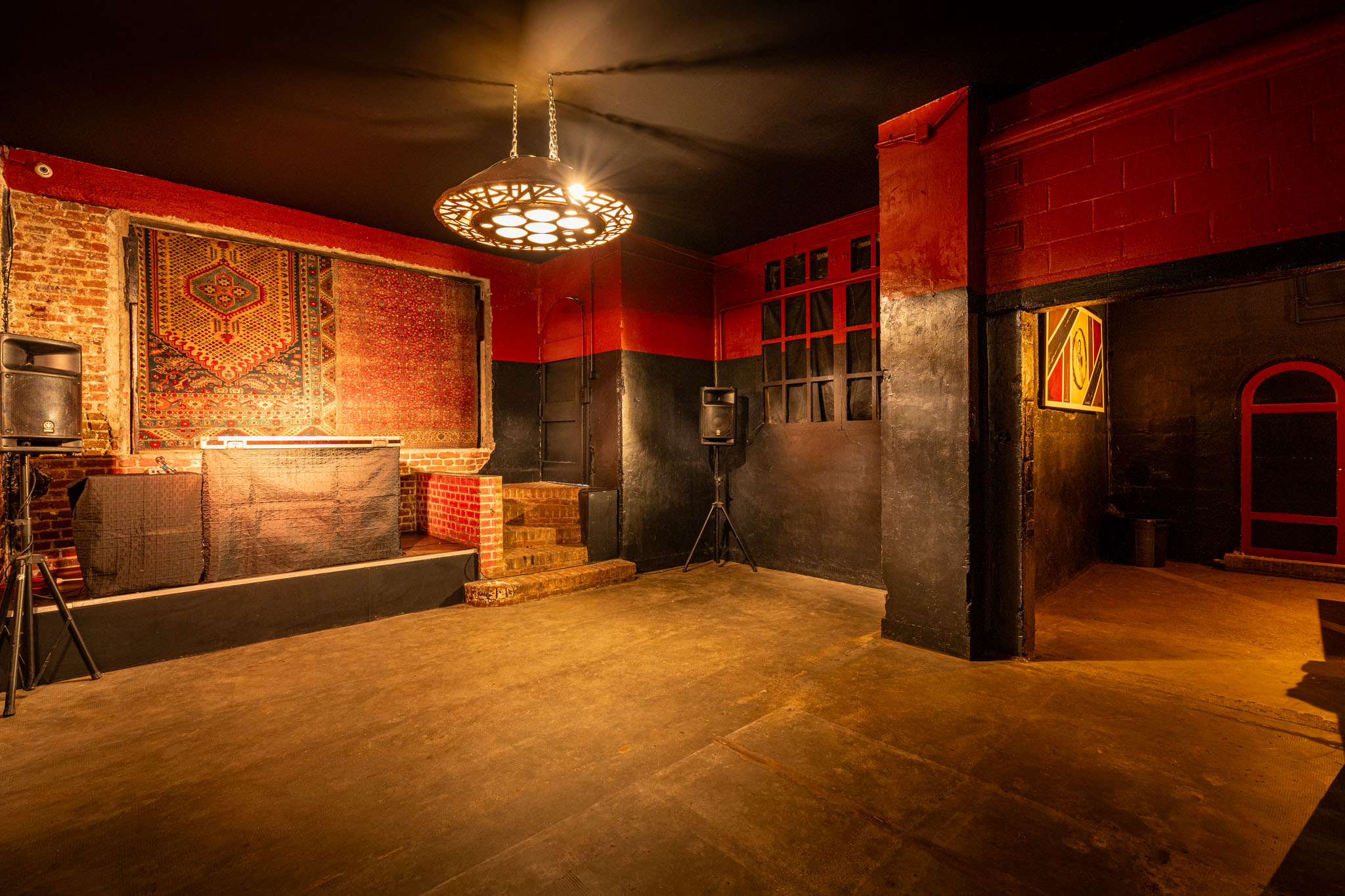 LA DEMEURE DU BOIS DE VERRIERES (92140) présente un club vide, faiblement éclairé, aux murs rouges et noirs, avec une cabine de DJ sur une petite scène, une lumière circulaire suspendue, deux haut-parleurs, une tapisserie murale et un sol en béton.