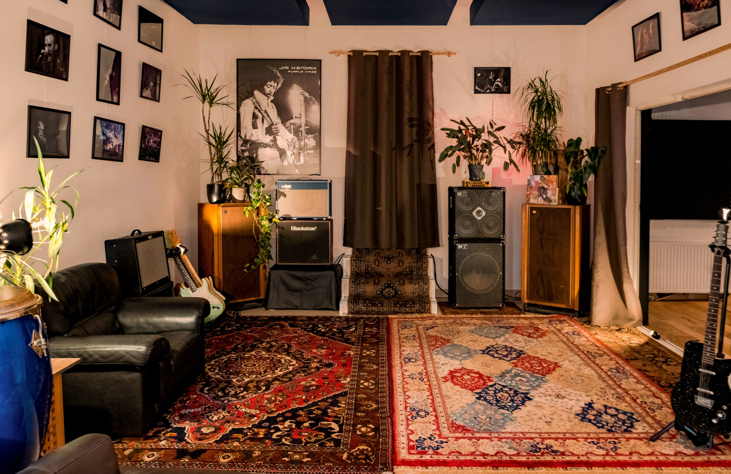 LA DEMEURE DU BOIS DE VERRIERES (92140) présente une salle de musique confortable avec des guitares, des amplis, des haut-parleurs, des photos de groupes encadrées, des plantes en pot, des tapis colorés, un canapé noir et des instruments disposés dans un éclairage chaleureux.
