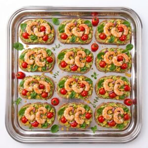 Un plateau en argent présente douze toasts de plantain à l’avocat et aux crevettes, chacun garni d’avocat, de crevettes, de légumes verts, de tomates cerises et d’herbes aromatiques, le tout disposé avec soin en trois rangées.