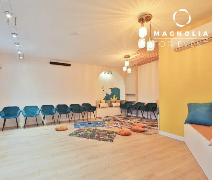 Une salle moderne et lumineuse à L'ESPACE DES AMIS DU 15ème (75015), avec un demi-cercle de chaises bleues, des tapis de jeu colorés, des coussins orange sur un sol en bois clair, des lumières douces au plafond et un mur d'accentuation jaune.