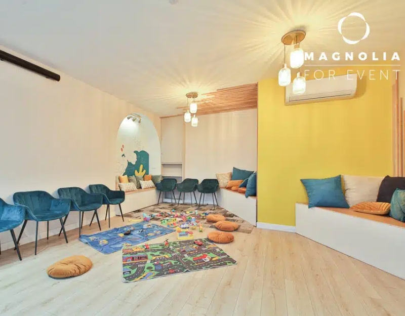Une salle de jeux moderne et lumineuse à L'ESPACE DES AMIS DU 15ème (75015) comprend des chaises bleues, des bancs muraux rembourrés, des tapis de jeu colorés avec des jouets, des planchers en bois, des murs jaunes et des lumières d'ambiance au plafond.