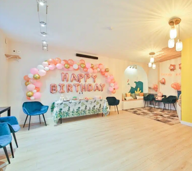 L'ESPACE DES AMIS DU 15ème (75015) présente une salle lumineuse décorée de ballons "Joyeux anniversaire", d'accents roses et dorés, d'une nappe fleurie sur la table et de plusieurs chaises bleues disposées dans l'espace.