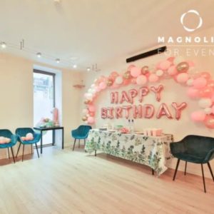 À L'ESPACE DES AMIS DU 15ème (75015), une table à la nappe fleurie est entourée de quatre chaises bleues, tandis que des ballons roses et blancs forment une arche au-dessus des ballons-lettres HAPPY BIRTHDAY pour une célébration festive.