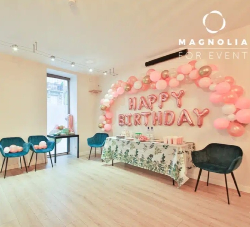 À L'ESPACE DES AMIS DU 15ème (75015), une table à la nappe fleurie est entourée de quatre chaises bleues, tandis que des ballons roses et blancs forment une arche au-dessus des ballons-lettres HAPPY BIRTHDAY pour une célébration festive.