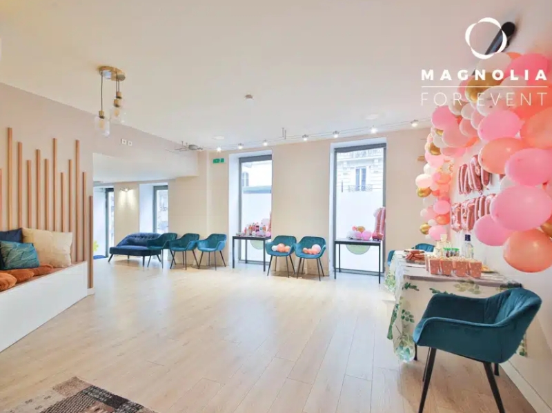 L'ESPACE DES AMIS DU 15ème (75015) dispose d'un espace événementiel lumineux avec des planchers en bois, des sièges pastel, de grandes fenêtres et une table de cadeaux avec des ballons roses HAPPY BIRTHDAY. La marque Magnolia For Event apparaît dans le coin.
