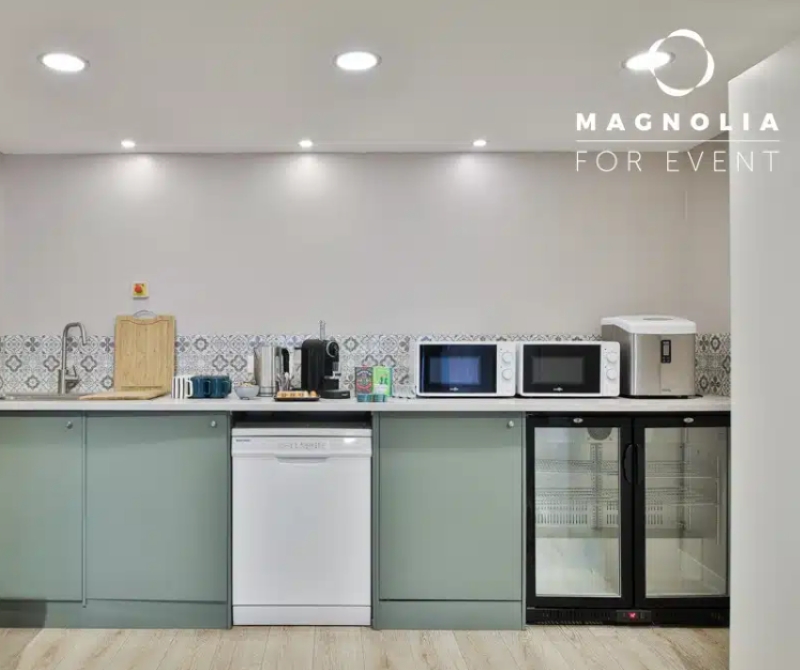 Cuisine moderne à L'ESPACE DES AMIS DU 15ème (75015) avec des armoires vert pâle, un évier, un lave-vaisselle, une cafetière, un micro-ondes, deux mini-réfrigérateurs à façade en verre. Le logo Magnolia For Event se trouve dans le coin supérieur droit.