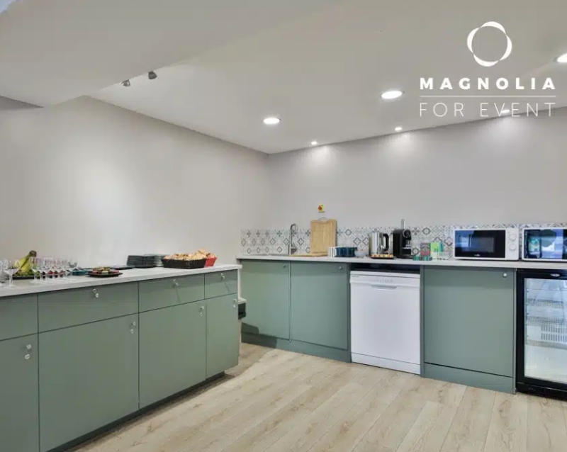 L'ESPACE DES AMIS DU 15ème (75015) dispose d'une kitchenette moderne avec des armoires vert sauge, un lave-vaisselle, un micro-ondes, une cafetière, des snacks sur le comptoir, un plancher en bois clair et un dosseret en carreaux à motifs. Le logo Magnolia For Event apparaît en haut à droite.