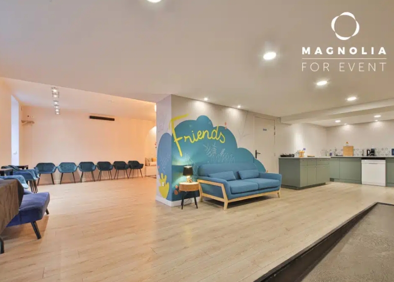 Espace événementiel moderne, L'ESPACE DES AMIS DU 15ème (75015), avec un sol en bois, un canapé bleu près d'un mur peint de Friends, des rangées de chaises bleues, une kitchenette, un éclairage lumineux et un logo dans le coin.