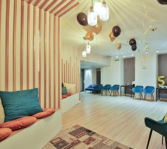 L'ESPACE DES AMIS DU 15ème (75015) dispose d'une salle moderne et lumineuse avec des ballons dorés et noirs, des lampes suspendues, des chaises bleues, des murs rayés, des sièges rembourrés, un tapis sur un sol en bois clair, et un ballon S doré sur la table du fond.