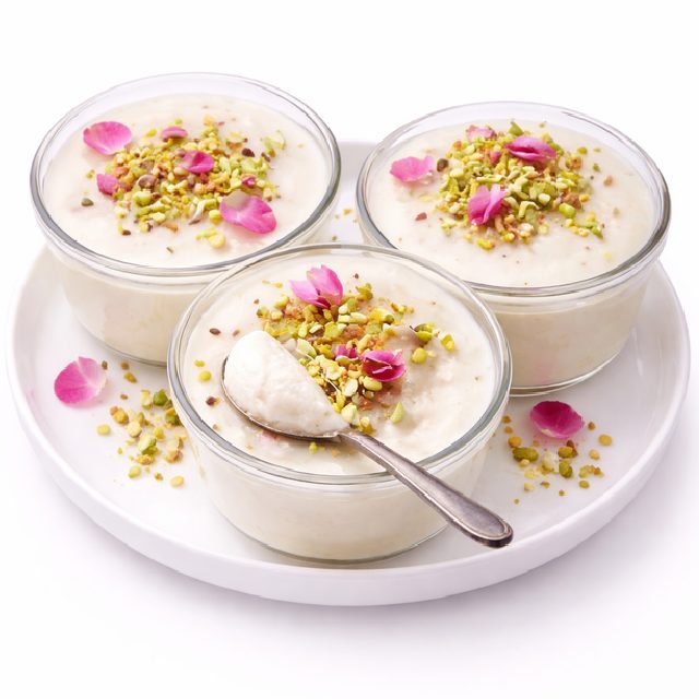 Trois coupes en verre de MOUHALLABIEH FLAN À LA FLEUR D'ORANGER, garnies de pistaches hachées et de pétales de roses, sont disposées sur une assiette blanche. Une cuillère est posée dans l'un des bols.