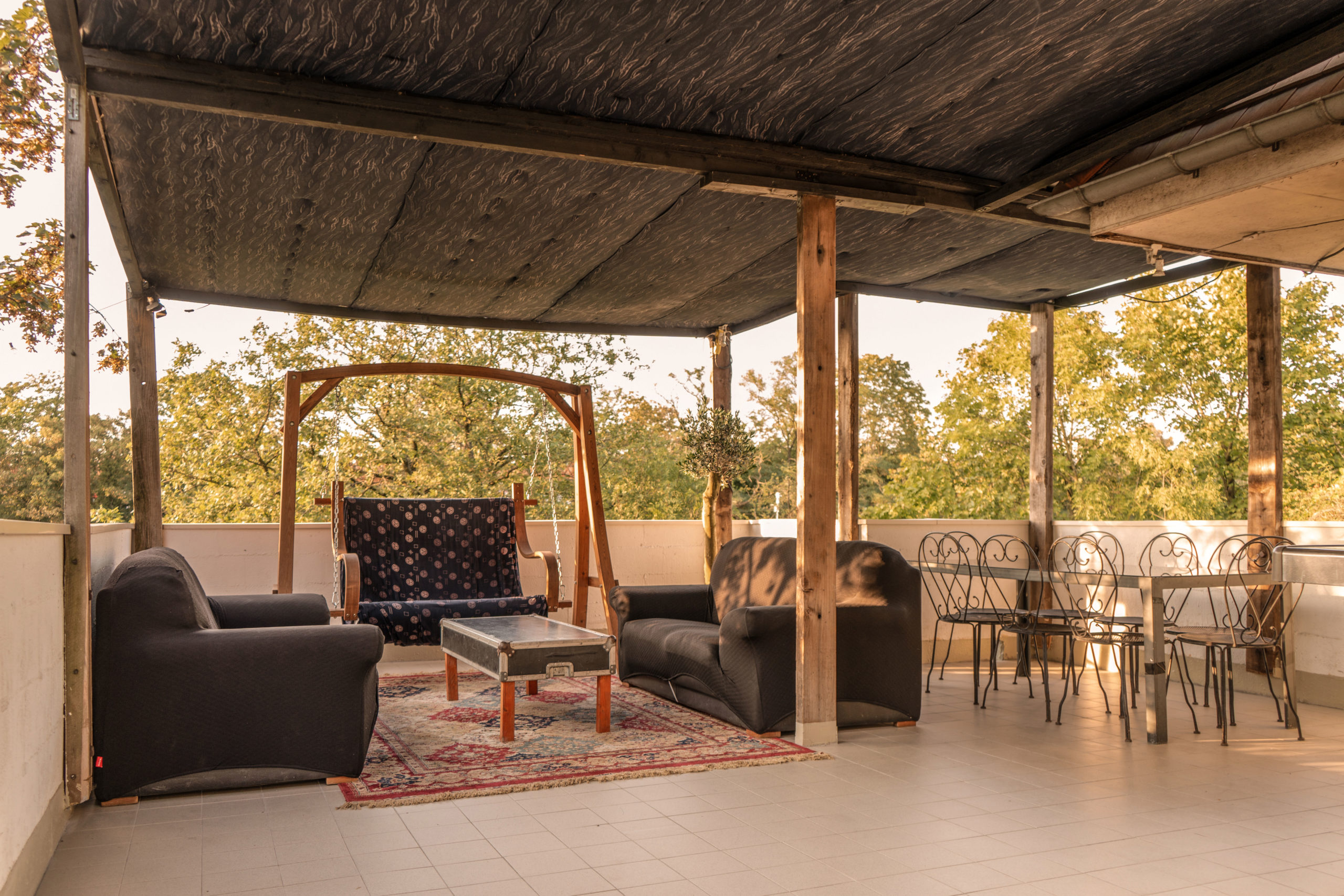 LA DEMEURE DU BOIS DE VERRIERES (92140) dispose d'une terrasse couverte spacieuse avec deux canapés noirs, une table basse recouverte d'un tapis, une balancelle en bois et une table de salle à manger en métal pour six personnes, le tout au milieu d'arbres et d'une lumière naturelle abondante.