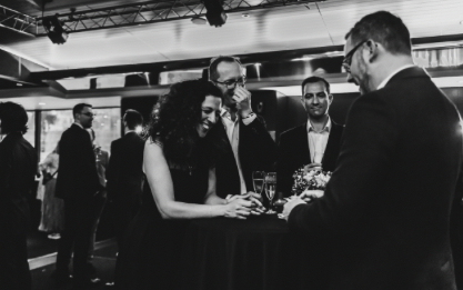 Les invités en tenue de soirée rient et se mêlent autour d'une table de cocktail sous un éclairage tamisé tandis qu'UN MAGICIEN PROFESSIONNEL livre un spectacle inoubliable, créant une atmosphère vivante et élégante lors de l'événement en salle.