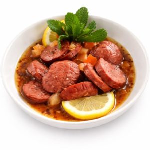 Un bol de SOUDJOUK SAUCISSES ÉPICÉES (Plat - Min de commande 5 plats de 300g/Pers), servi dans une sauce brune avec des tranches de citron, des dés de légumes et un brin de menthe fraîche sur le dessus.