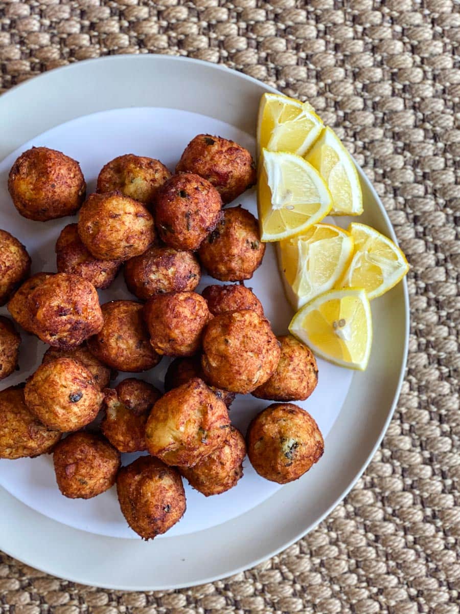 Une assiette d'ATELIER ACRAS DE MORUE À VOLONTÉ, boules de morue frites et dorées, est disposée avec des quartiers de citron sur une assiette blanche et beige surmontant un napperon tissé.