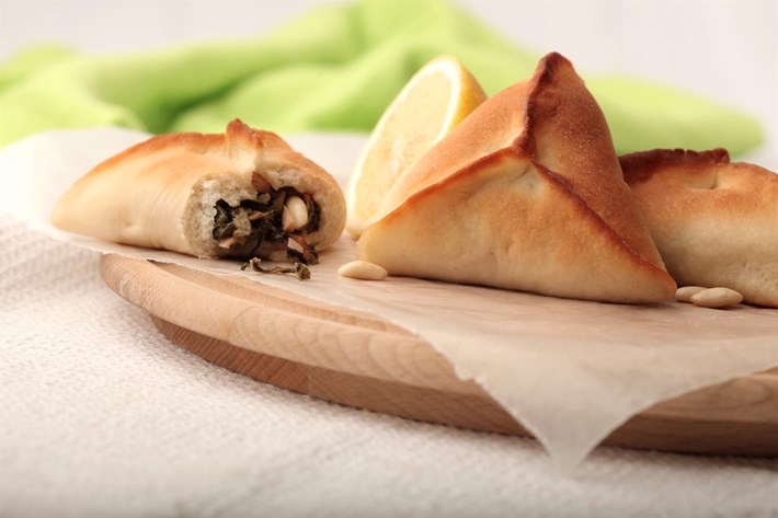 Trois FATAYER ÉPINARDS (80 g chacun), dont l'un est ouvert pour révéler une garniture d'épinards et de pignons, reposent sur une planche en bois parcheminée, avec un quartier de citron en arrière-plan. Commande minimum : 5 pièces. Entrée chaude.