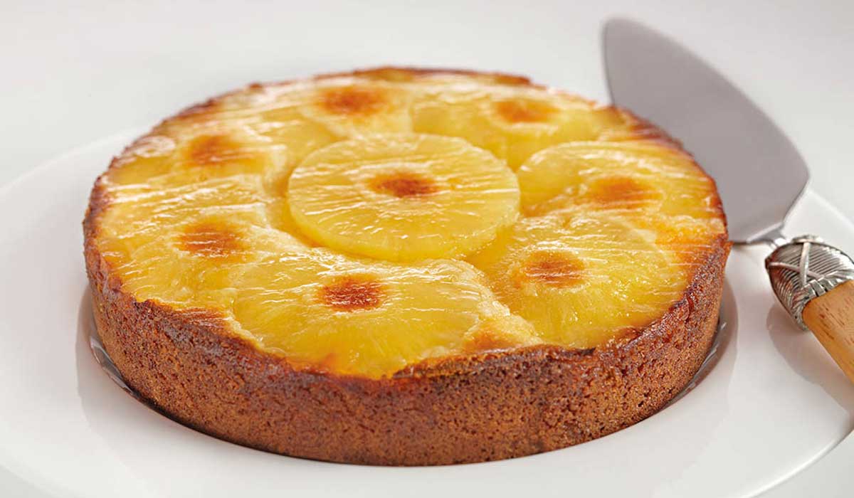 Un gâteau renversé à l'ananas, garni de tranches d'ananas disposées en cercle et aux bords dorés, trône sur une assiette blanche, à côté d'une pelle à gâteau en métal munie d'un manche en bois.