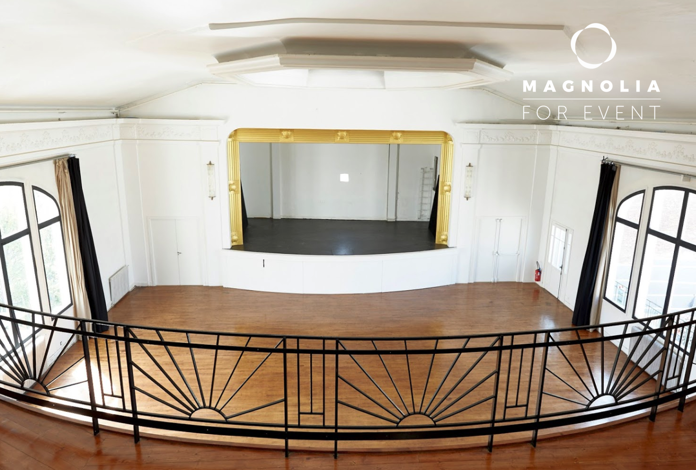 Salle d'événements spacieuse et vide avec des planchers en bois, une petite scène encadrée d'or, de grandes fenêtres et une balustrade noire de style art déco. L'inscription "LE LOFT SOLARIS (75020)" figure dans le coin supérieur droit.
