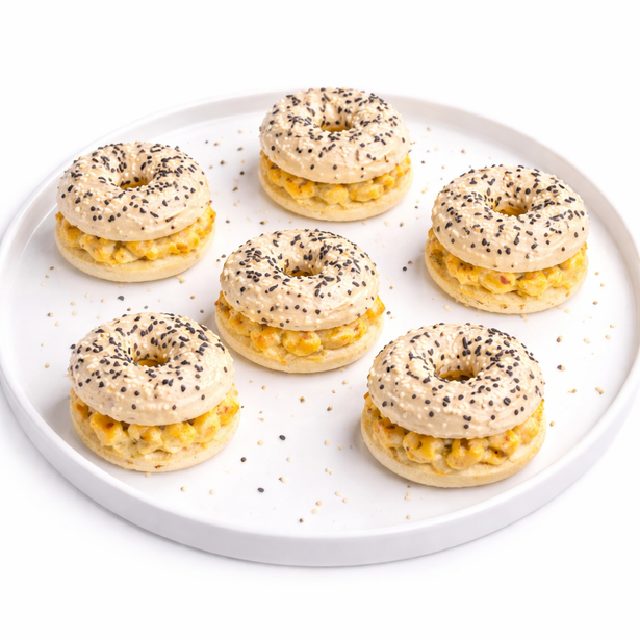 Dix MINI BAGELS POULET CURRY (20 g chacun, commande minimum) sont disposés sur une assiette ronde blanche. Les mini-bagels sont fourrés au poulet au curry et recouverts de graines de pavot et de sésame. Quelques miettes sont éparpillées sur l'assiette.