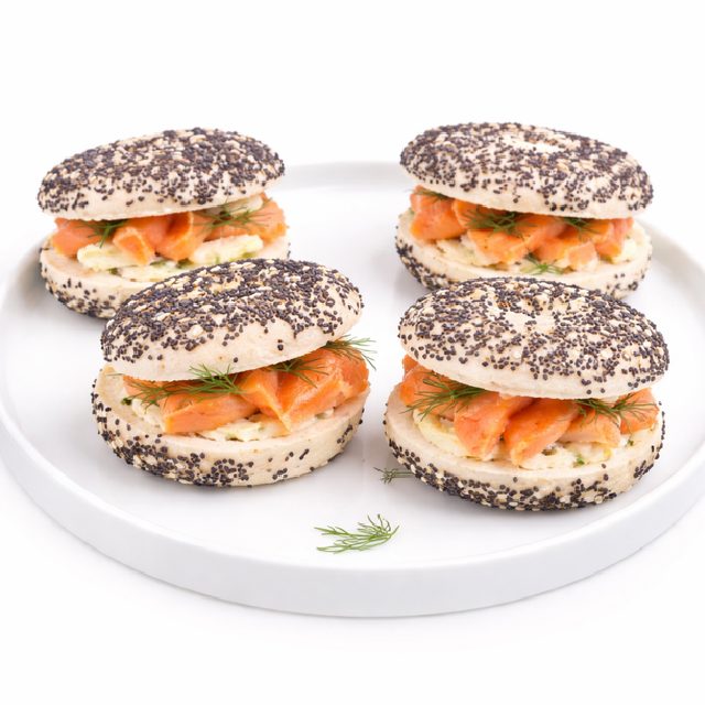 Les MINI BAGELS SAUMON (20g/pièce, minimum 10) sont fourrés de saumon fumé, de fromage frais et d'aneth frais, servis dans une assiette ronde blanche et garnis d'aneth supplémentaire.