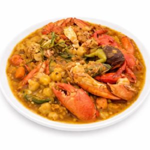 Une assiette blanche présente le DOMBRÉ DE CRABES EN COLOMBO (300 g), un ragoût de fruits de mer composé de pinces de homard, de pois chiches, de légumes et d'une riche sauce dorée. Ce plat copieux et coloré se détache sur le fond blanc. Commande minimum : 5 plats.